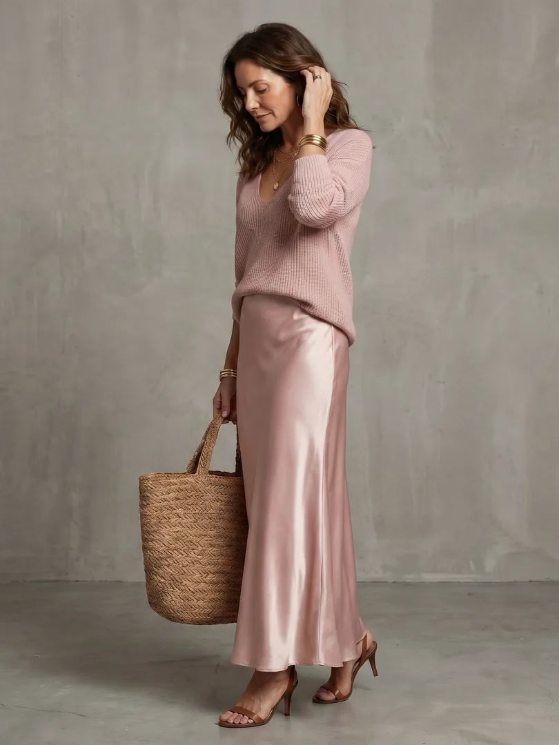 Side profile in pink knit jumper and satin bias skirt, adjusting hair, holding a woven straw tote bag
