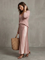 Side profile in pink knit jumper and satin bias skirt, adjusting hair, holding a woven straw tote bag