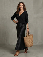 Full front view in black ribbed V-neck jumper paired with a black satin bias-cut midi skirt and straw tote