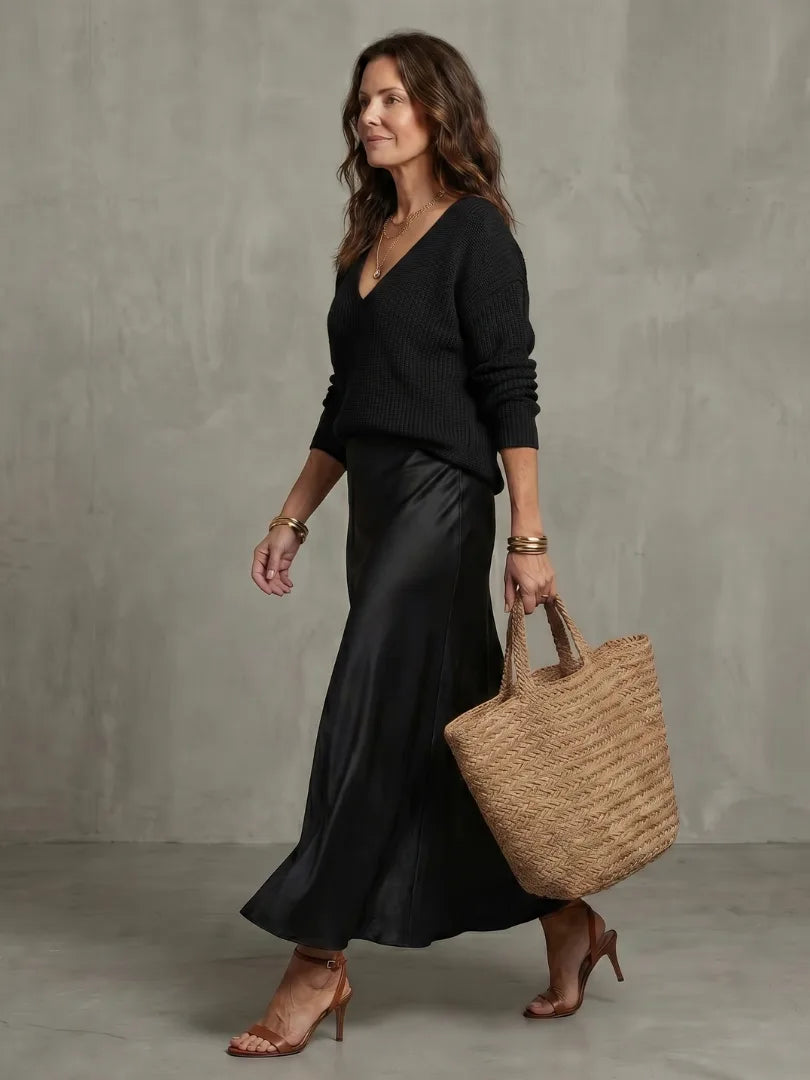Side walking shot in black knit jumper and black satin skirt with woven tote bag and brown heeled sandals