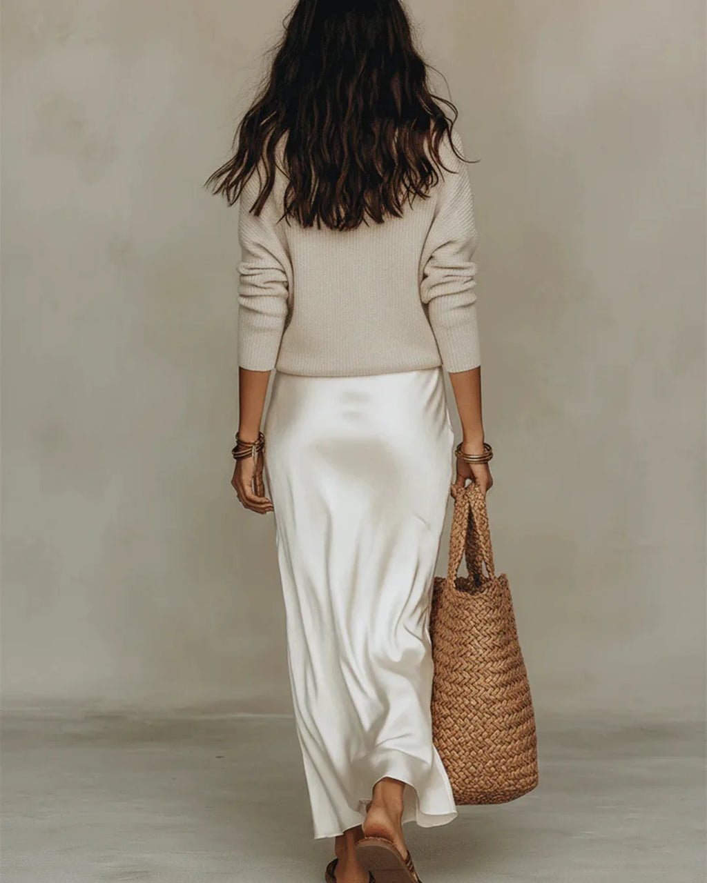 Back view of a model in a cream jumper and flowing ivory satin midi skirt, walking away with a straw tote bag