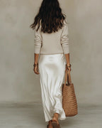 Back view of a model in a cream jumper and flowing ivory satin midi skirt, walking away with a straw tote bag