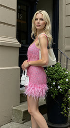 Side profile of a fringe sequin mini dress in pink with a feather hem, worn with a white shoulder bag on a doorstep