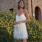 Smiling woman wearing a white sequin fringe mini dress with feather trim, holding a white top-handle bag outdoors