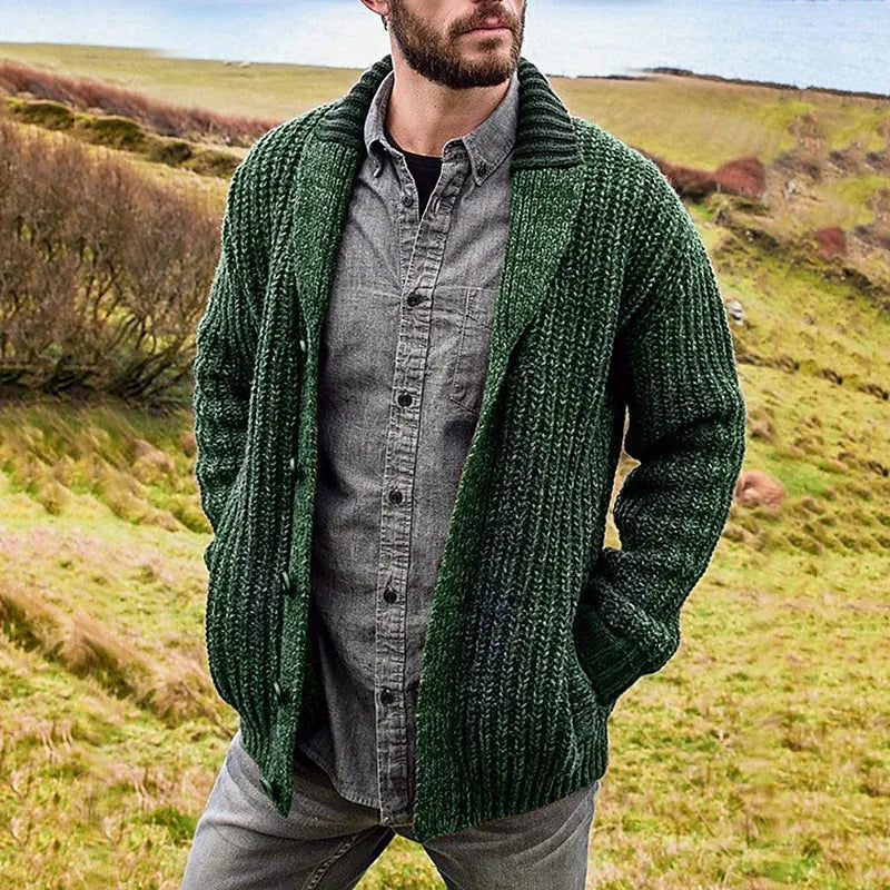 Close view of a green textured knit cardigan with shawl collar and open front, worn over a grey shirt in a countryside field