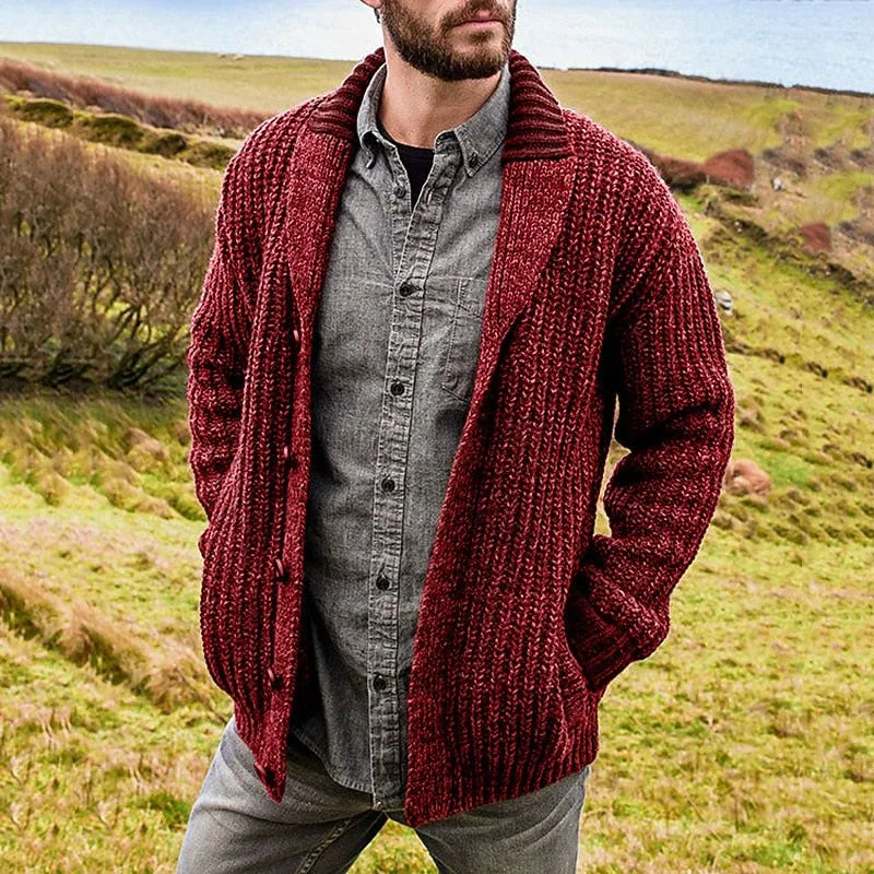 Man modelling a burgundy red knit shawl collar cardigan with visible ribbed cuffs and pockets, standing in a grassy coastal landscape
