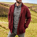 Man modelling a burgundy red knit shawl collar cardigan with visible ribbed cuffs and pockets, standing in a grassy coastal landscape