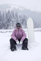 Side view of the same dusky pink hooded ski jacket paired with black salopettes, seated in snowfall
