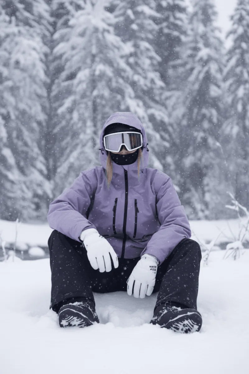 Front view of a lilac purple hooded ski jacket worn with a white snow goggle and gloves, standing among snowy pines
