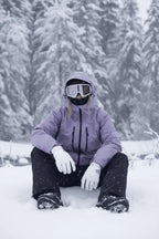 Front view of a lilac purple hooded ski jacket worn with a white snow goggle and gloves, standing among snowy pines