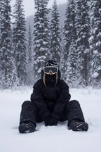 Head-to-toe all black snowsuit with hood, goggles and matching beanie, seated in falling snow