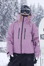 Close-up front detail of the dusky pink jacket showing zip chest pockets and storm hood, standing beside a snowboard