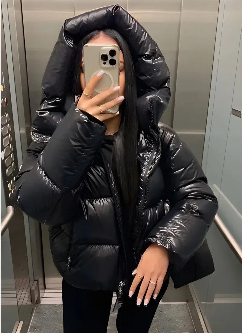 Woman wearing a black glossy puffer jacket with oversized hood pulled up, taking a mirror selfie in a lift