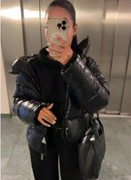 Woman wearing a black glossy puffer jacket with hood down, zipped up over a black outfit, mirror selfie