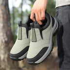 Grey and black slip-on outdoor trainer held in hand, showing textured upper and chunky lugged sole against a forest backdrop