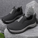 Black slip-on outdoor trainer with grey ridged sole resting on rocks, side view showing reinforced toe and pull tab
