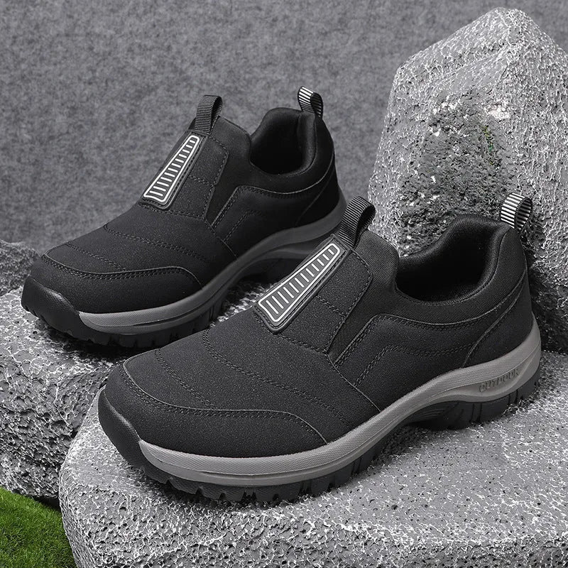 Black slip-on outdoor trainer with grey ridged sole resting on rocks, side view showing reinforced toe and pull tab