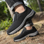 Dark blue suede-effect slip-on trainer with black overlays and grey ridged sole, side profile on rock and grass background