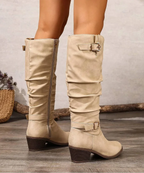 Back view of slouched beige knee-high boots with gold buckle straps and a stacked block heel, worn with bare legs