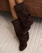 Overhead shot of a woman seated wearing dark brown suede slouchy boots with ruched folds, feet crossed on a rug