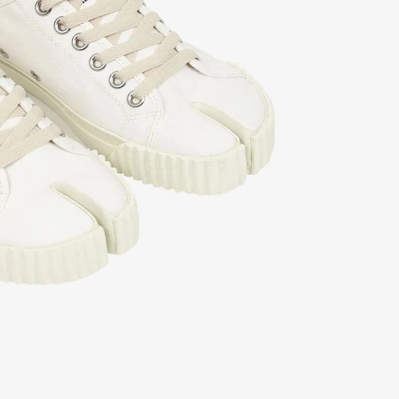 Cropped view of a person's ankles wearing white split-toe canvas trainers with a grey midi skirt