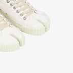 Cropped view of a person's ankles wearing white split-toe canvas trainers with a grey midi skirt