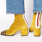 Back view of mustard yellow suede ankle boot with leopard-print toe cap and red zip trim, worn with jeans