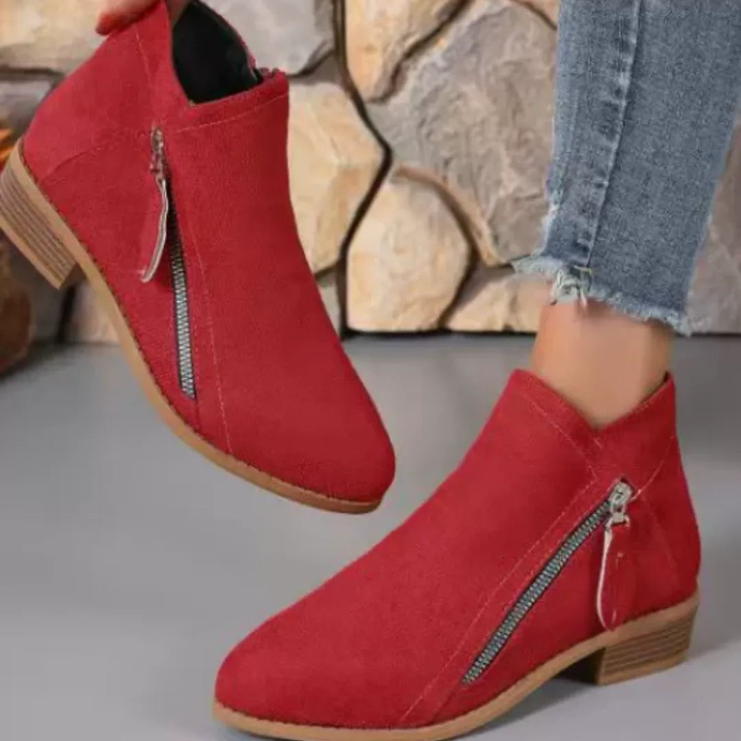 Red suede ankle boots worn against a stone wall backdrop, showing side zipper and tan block heel