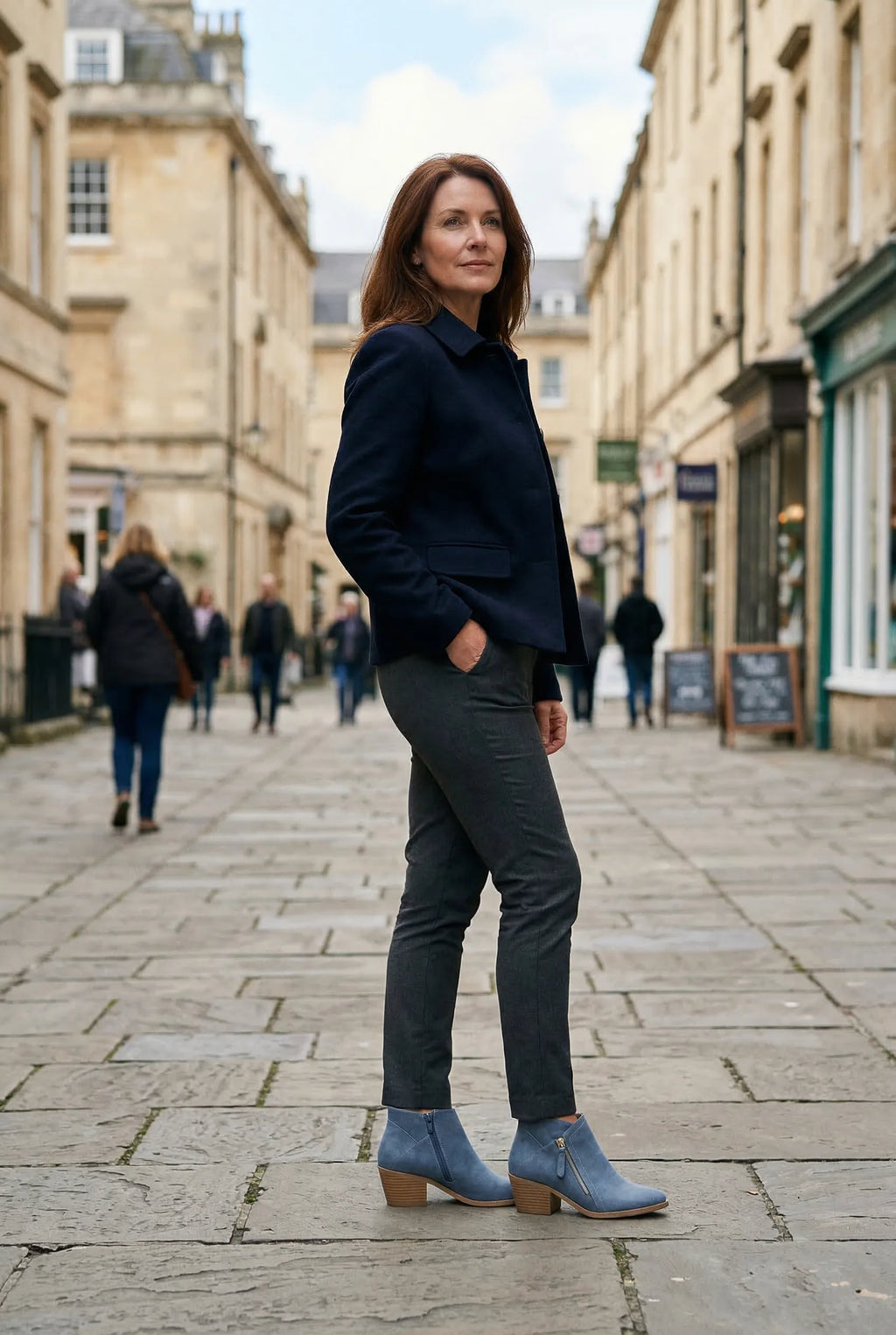 Woman in a navy blazer and dark trousers walking a cobbled street wearing blue ankle boots