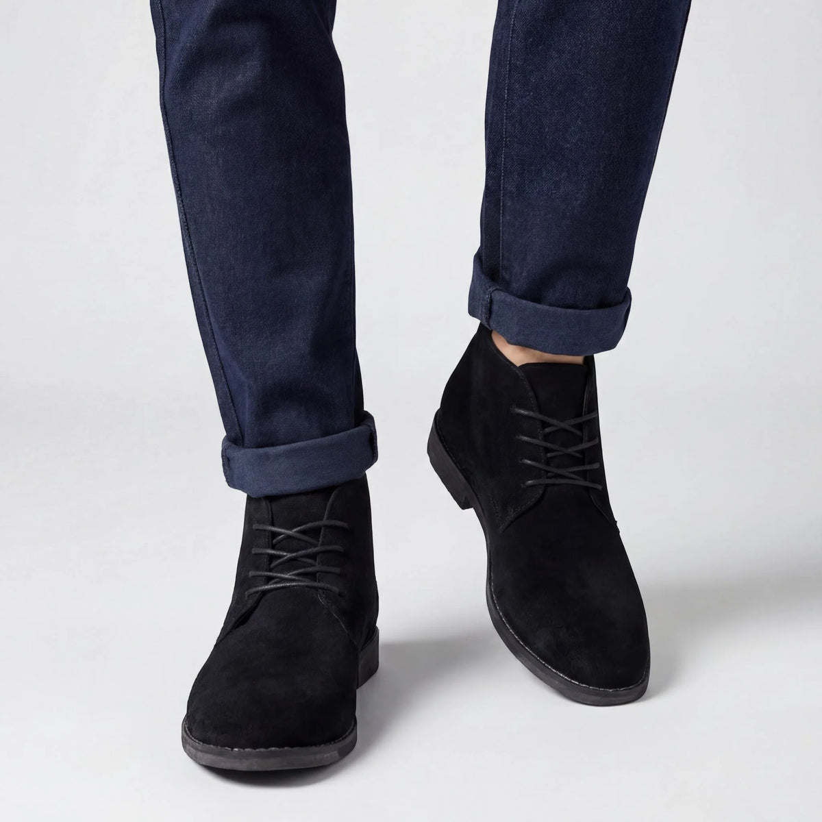 Close-up front view of black suede chukka boots worn with rolled navy jeans, laced front detail visible