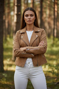 Front view of a tan suede-effect biker jacket worn over a white top and white jeans, arms crossed showing zip cuffs.
