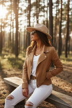 Side view of the tan biker jacket paired with a hat, sunglasses and layered necklaces, seated on a wooden bench.