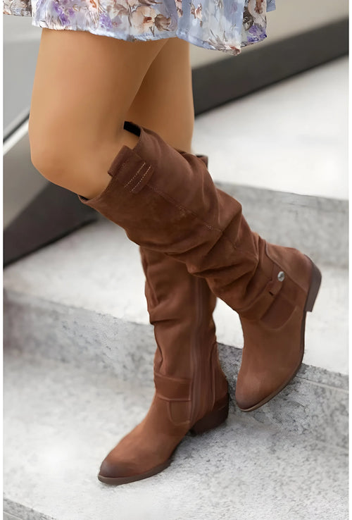 Side view of tan suede knee-high boots with buckle strap detail and slouch fold, worn with a floral dress on steps