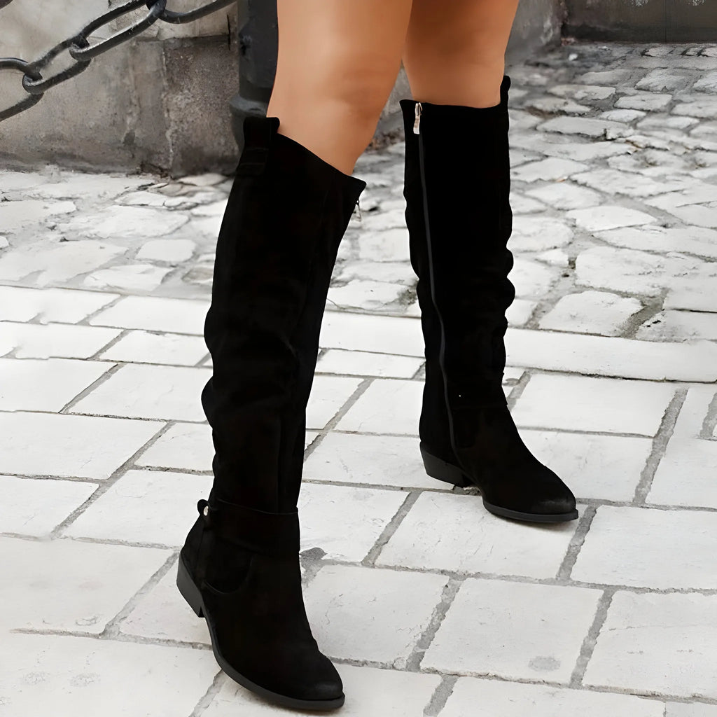 Black suede knee-high boots with side zip and pointed toe, worn walking on a cobbled street