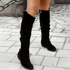 Black suede knee-high boots with side zip and pointed toe, worn walking on a cobbled street