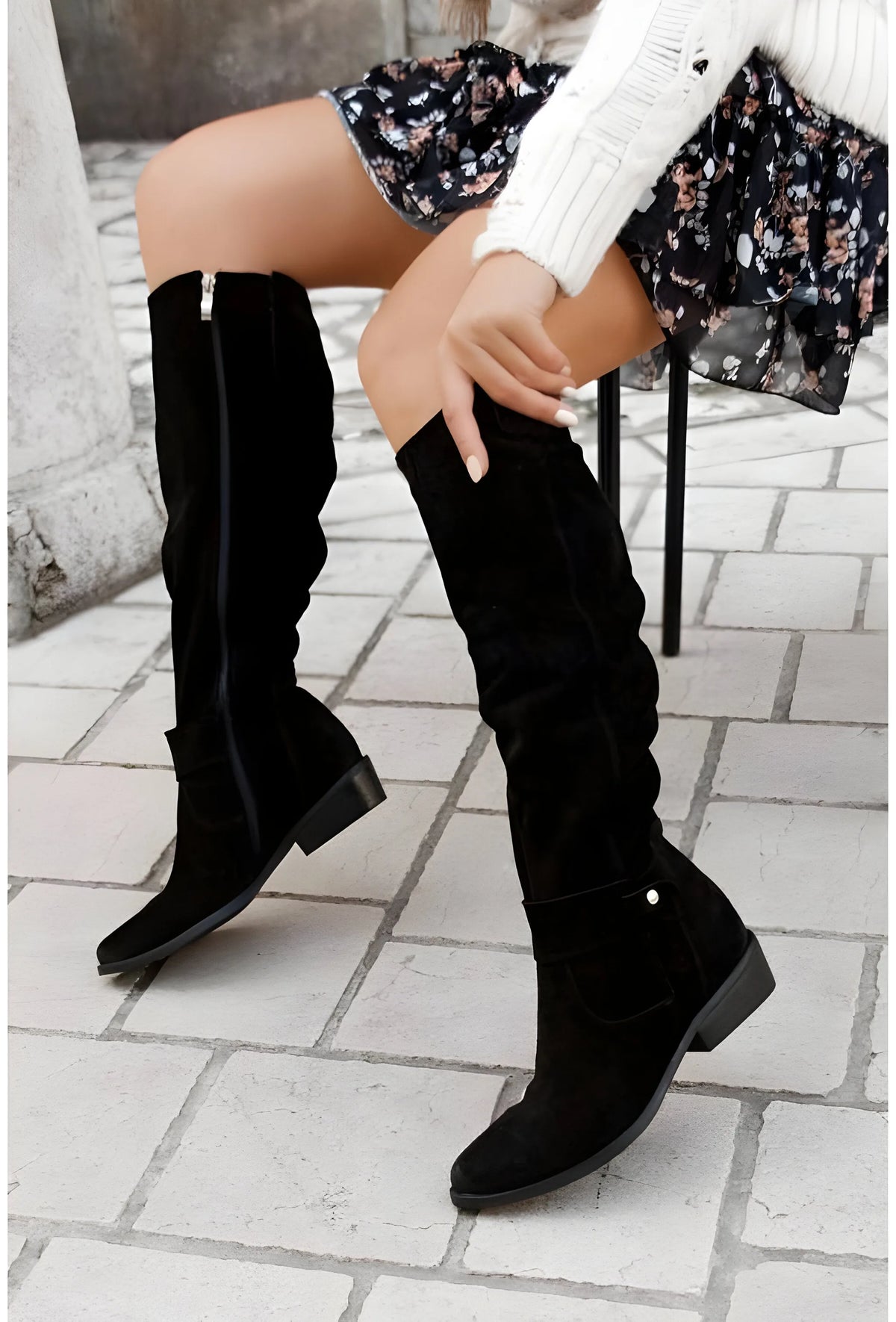 Seated view of black suede knee-high boots with slouch folds, ankle strap and stacked heel, worn with a floral skirt