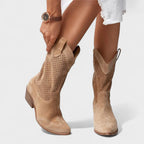 Woman adjusting a tan suede cowboy-style boot, showing pointed toe and perforated side panel texture