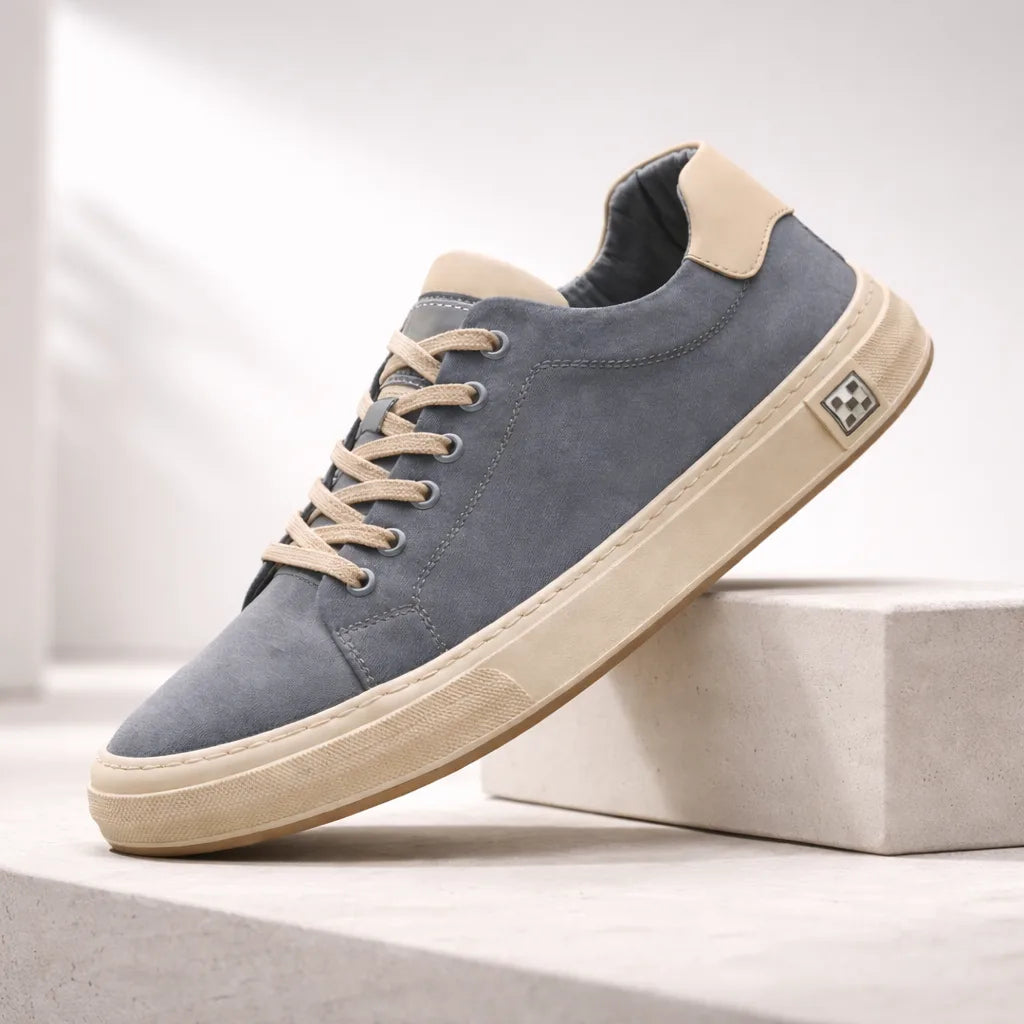 Angled shot of a blue suede trainer resting against a concrete block, showing stitched toe cap and beige heel panel