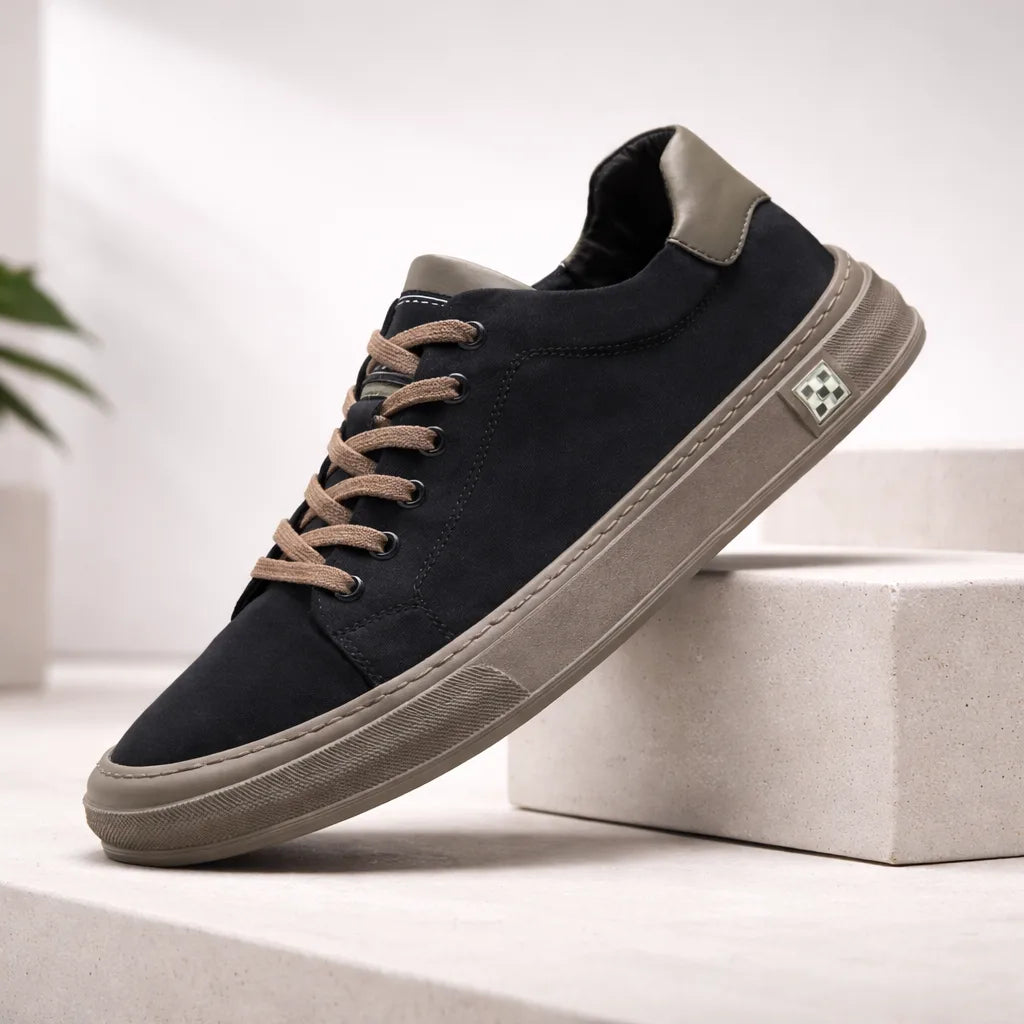 Black suede low-top trainer propped on a concrete block, featuring tan laces and a grey heel panel