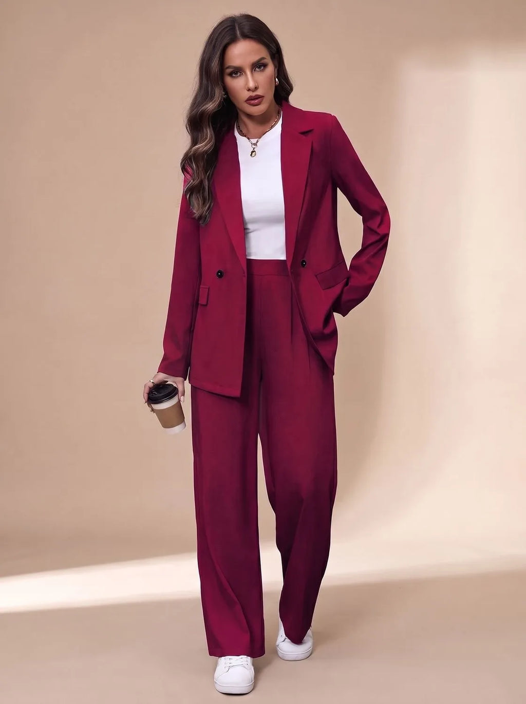 Front view of the burgundy tailored trouser suit styled with a white top and white trainers, holding a coffee cup