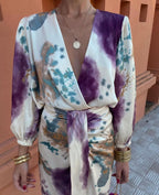 Close-up of a tie-dye print wrap dress in ivory, purple and teal with a deep V-neckline and gold layered necklace