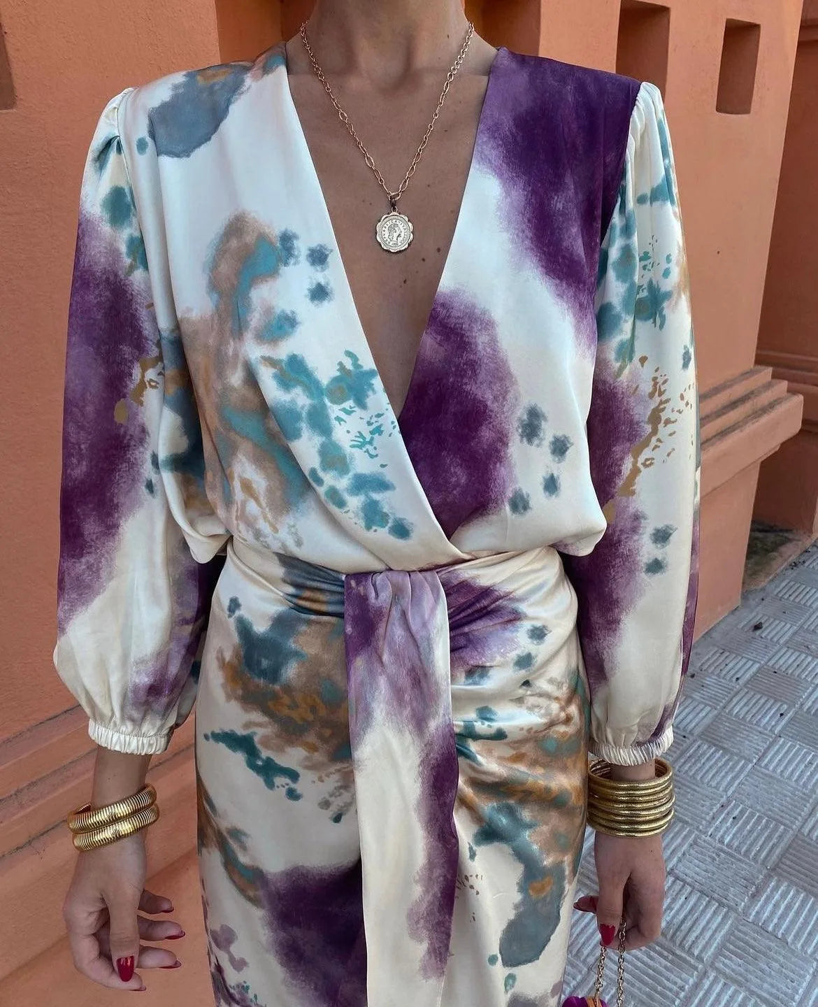 Close-up of a tie-dye print wrap dress in ivory, purple and teal with a deep V-neckline and gold layered necklace