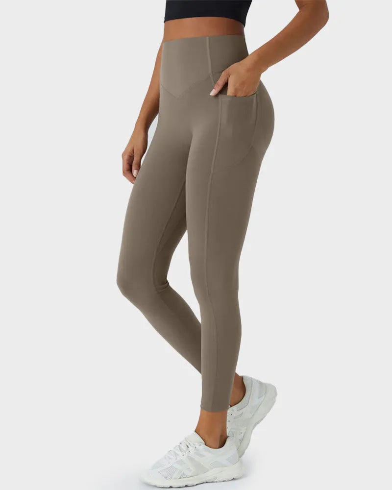Side view of high-waisted taupe leggings with hand tucked into deep side pocket, worn with a black crop top and white trainers