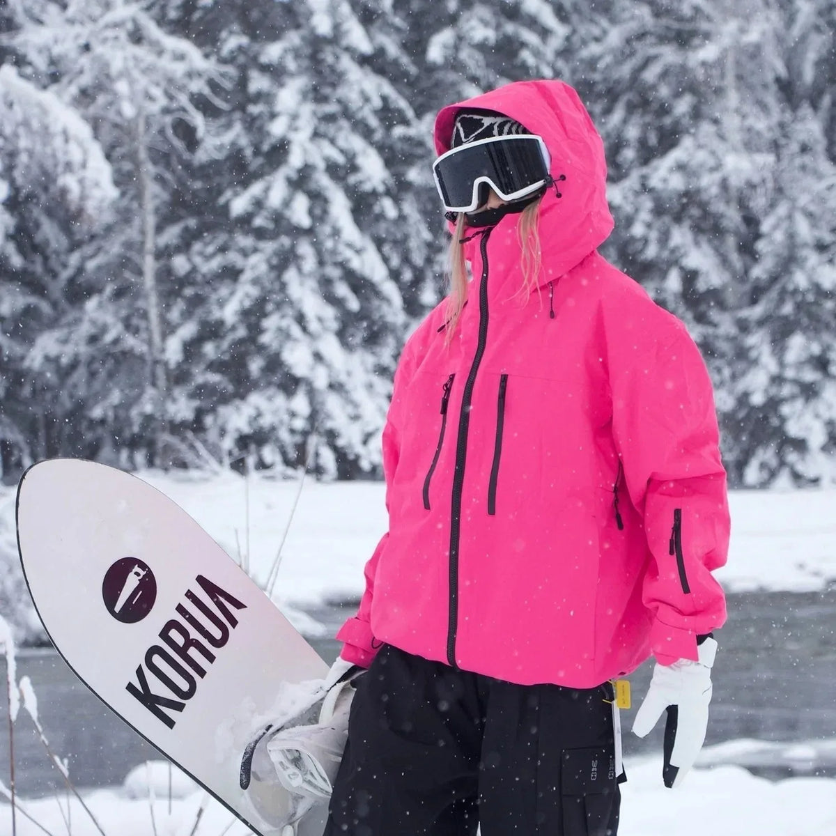 Model in a bright pink ski jacket with hood up, holding a white snowboard in snowy woodland