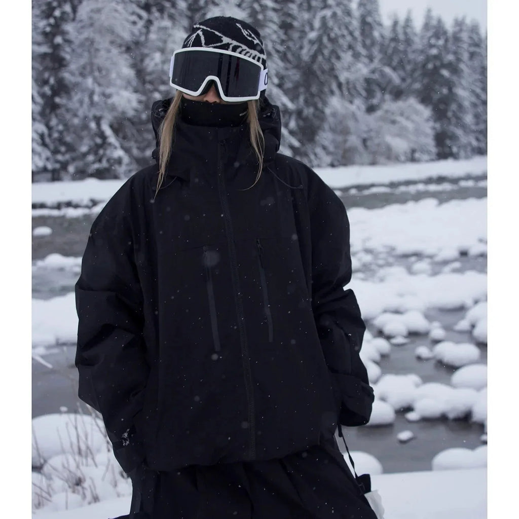 Model wearing an all-black ski jacket with hood up, standing by a snowy river bank