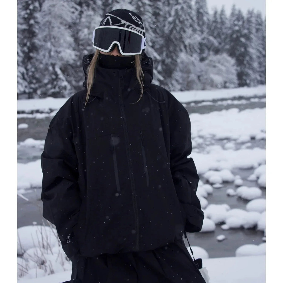 Model wearing an all-black ski jacket with hood up, standing by a snowy river bank