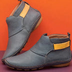 Slate blue leather ankle boots with mustard side strap and ridged sole, shown against an orange backdrop