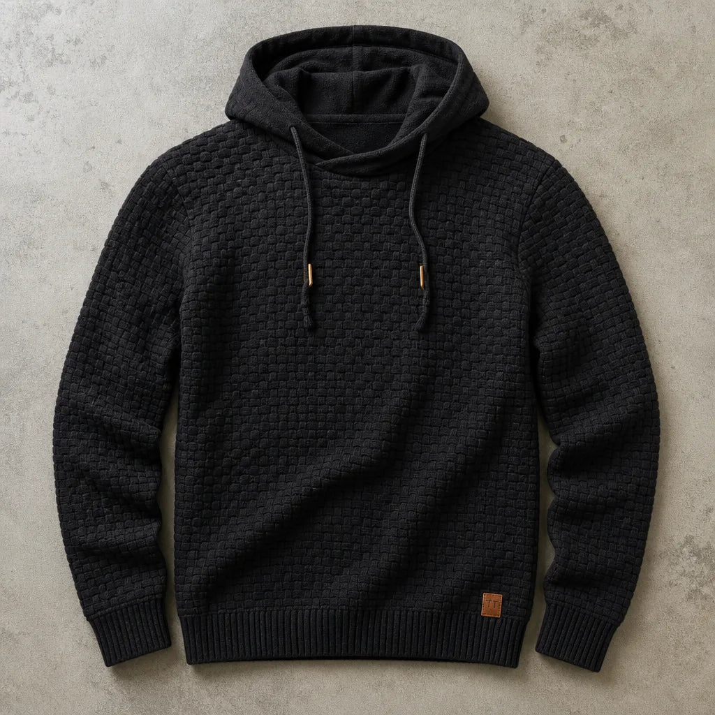 Black waffle-knit cowl-neck pullover hoodie laid flat, showing textured basket-weave knit and metal-tipped drawstrings