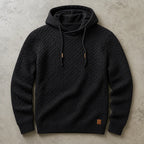 Black waffle-knit cowl-neck pullover hoodie laid flat, showing textured basket-weave knit and metal-tipped drawstrings