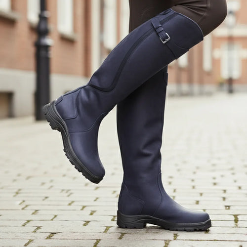 Side view of navy blue knee high leather boot with buckle strap, lugged sole raised mid-stride on cobbles